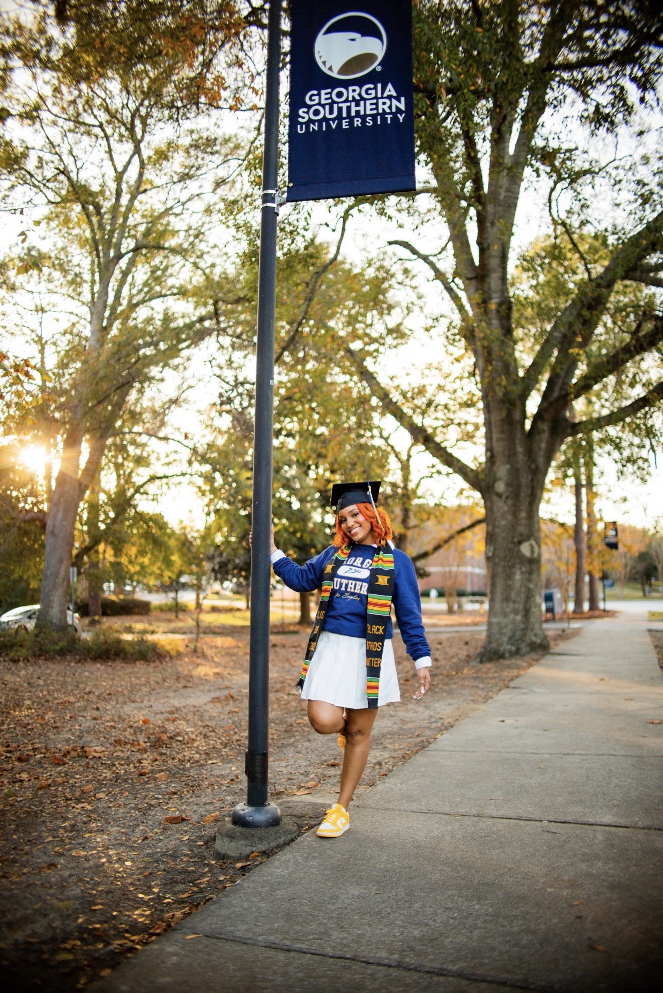 Taylor Pledger in a white skirt, blue Georgia Southern sweatshirt, yellow sneakers, graduation cap and stole. She is holding a pole with a blue Georgia Southern banner. Photo Credit: Gregory Martin