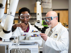 Student in lab coat experiencing hands on training with professor and high tech machinery.