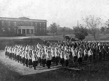 A black-and-white photo shows a large group of students in uniform standing in rows outside a brick school building, with several adults nearby and an early automobile in the background.