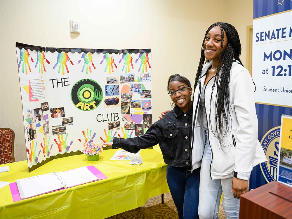 Two smiling students stand together beside a colorful Art Club display board on a table with yellow cloth at a school event. The board features photos, handprints, and the words The Art Club in bold letters.
