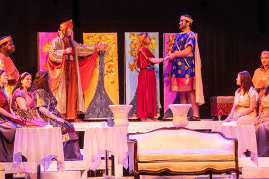 A theatrical performance with actors in colorful, ancient-style costumes on stage. Two main actors stand and hold hands while others sit around tables, with painted trees as the backdrop.