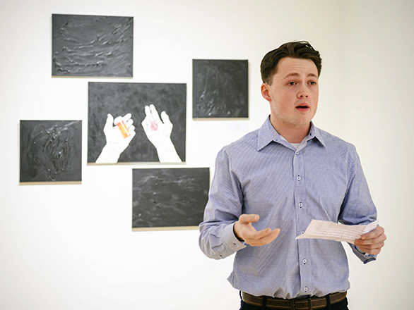 A young man in a blue shirt speaks while holding papers, standing in front of a wall display featuring black panels and a set of hands holding objects.
