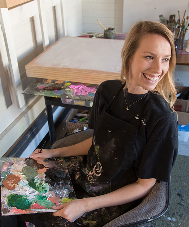 A smiling woman wearing a paint-stained apron sits in an art studio, holding a palette covered in mixed paint colors, with a blank canvas and art supplies in the background.
