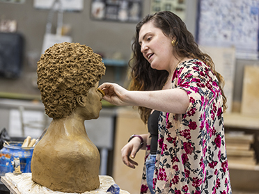 A woman in a floral kimono shapes the nose of a clay bust in an art studio, focusing intently on her sculpting work. Art supplies and tools are visible in the background.