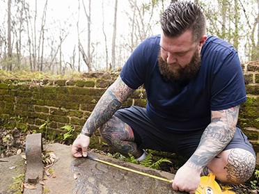 A man with tattoos and a beard kneels outdoors, measuring a stone slab with a yellow tape measure. He is wearing a navy blue shirt and shorts, next to a mossy brick wall in a wooded area.