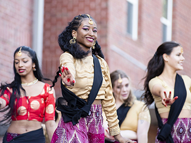 Four women dressed in colorful traditional outfits perform a dance outdoors, smiling and extending their arms, with a brick building in the background.