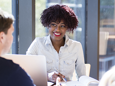 A woman with curly hair and a light denim shirt smiles while talking to another person across a table, with a laptop and notebook in front of her in a bright, modern office setting.