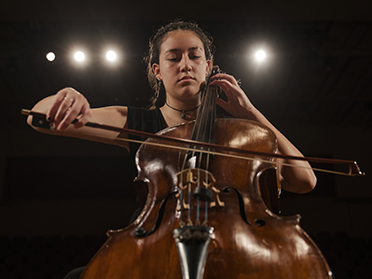 A young woman with curly hair plays the cello on stage, eyes closed in concentration, with bright stage lights shining behind her in a darkened auditorium.