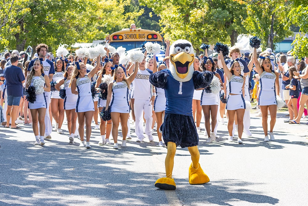 Gus in a number one jersey leads a group of cheerleaders in a parade. The cheerleaders, wearing blue and white Georgia Southern uniforms and waving pom-poms, march enthusiastically down a sunny street. A school bus and a crowd of spectators can be seen in the background under green trees.