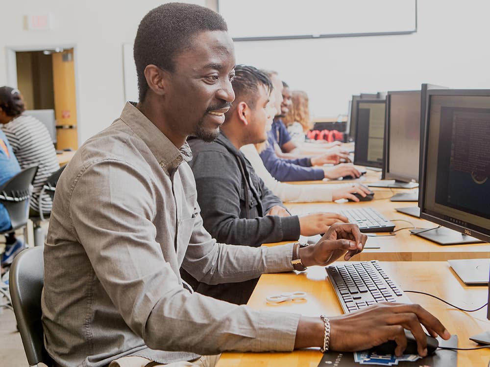 Young man working at a computer in a classroom full of other students learning information systems and analytics