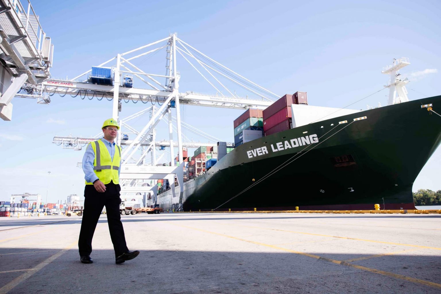 Parker College of Business logistics student in safety gear walking near cargo ship at a busy port terminal.