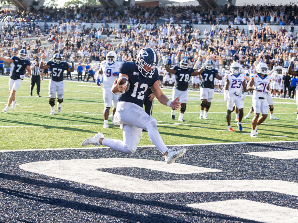A football player in a navy blue uniform leaps into the end zone to score a touchdown, while teammates and opponents look on and the crowd cheers in the background.