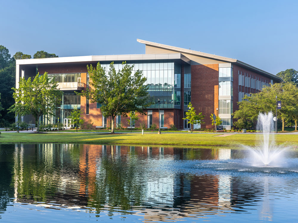 A modern, multi-story building with large glass windows is reflected in a calm pond with a water fountain, surrounded by green trees and a clear blue sky.