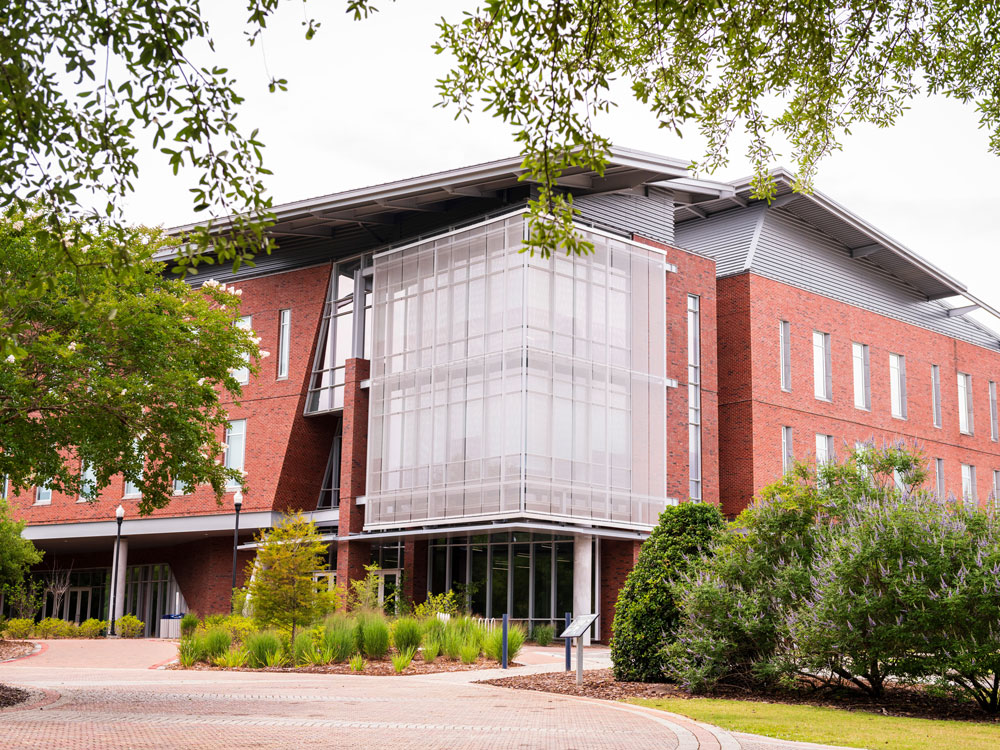A modern brick and glass building with three stories, surrounded by green trees and landscaped bushes, under a partly cloudy sky.