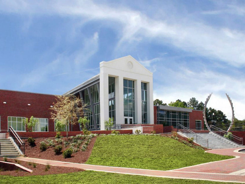 A modern red-brick building with large glass windows and white columns, surrounded by landscaped greenery and steps, under a blue sky with wispy clouds.