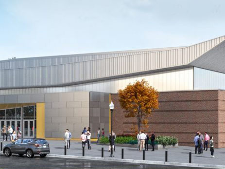 Several people stand and walk outside a modern building with large windows, beige and brick walls, and a tree with orange leaves near the entrance. A parked car is visible on the street in front.