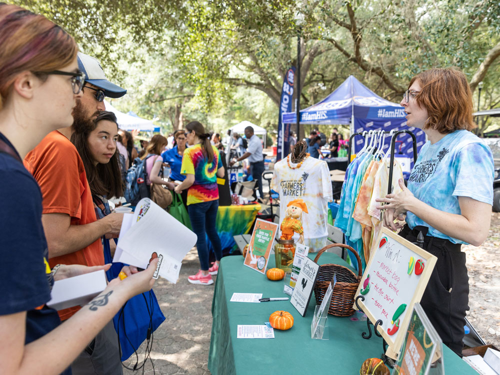 A group of people listens to a vendor at an outdoor farmers market booth. The booth displays colorful shirts, signs, and small pumpkins, with trees and other booths visible in the background.