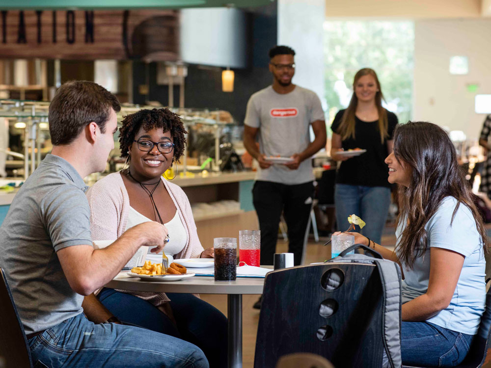 Three people sit at a table eating and talking in a bright cafeteria, while three others stand in the background carrying trays and smiling as they approach the table.
