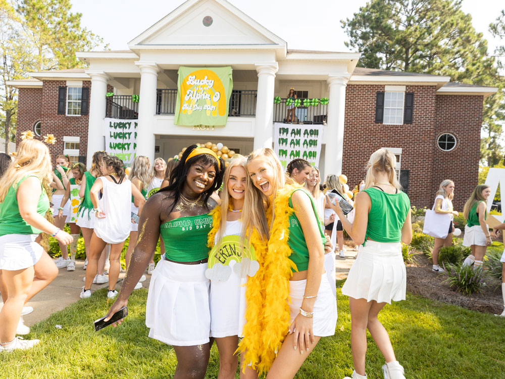 Three smiling young women pose together outside a sorority house during an event. They wear green and white outfits with accessories like a yellow feather boa. Other people and decorations are visible in the background.