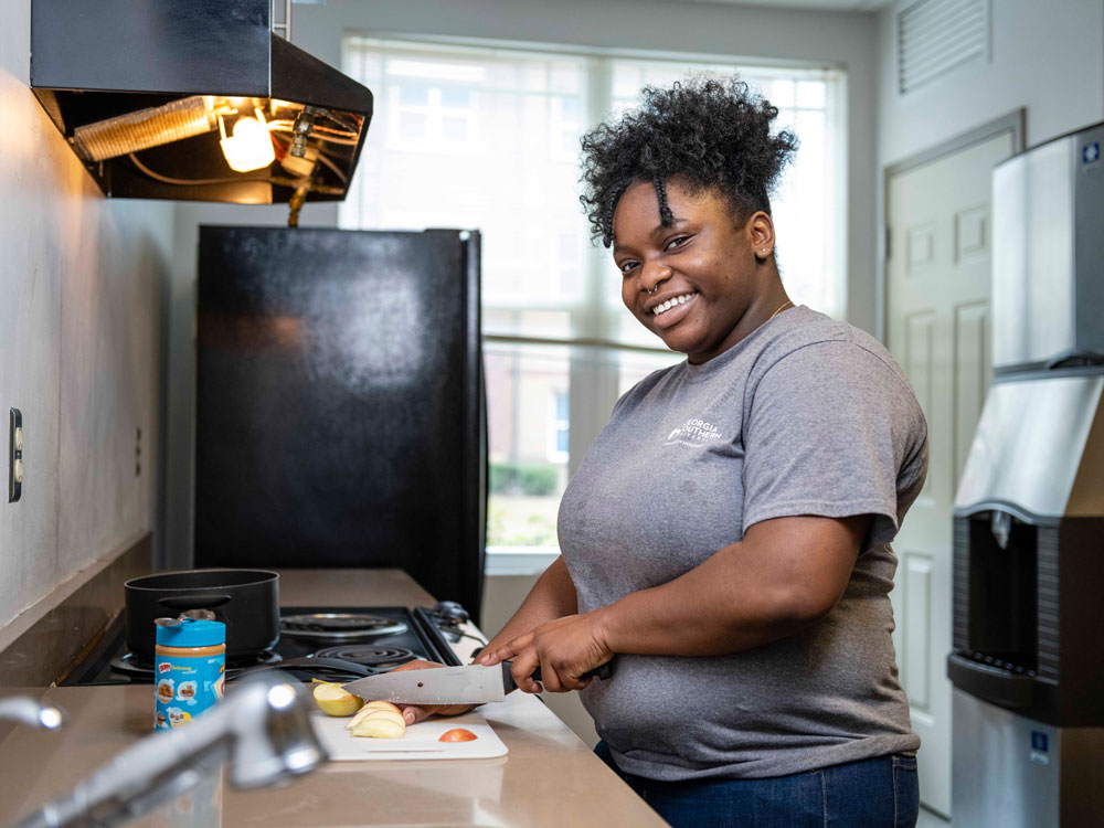A woman smiles while chopping vegetables on a cutting board in a modern kitchen. She stands near a stove and a window, wearing a gray shirt and jeans.
