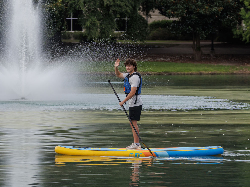 A person wearing a life jacket stands on a paddleboard, holding a paddle and waving, on a calm lake with a fountain spraying water in the background. Trees and grass line the shore.