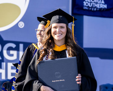 A smiling graduate in a cap and gown holds a diploma during a commencement ceremony at Georgia Southern University, with officials in academic regalia standing behind her.