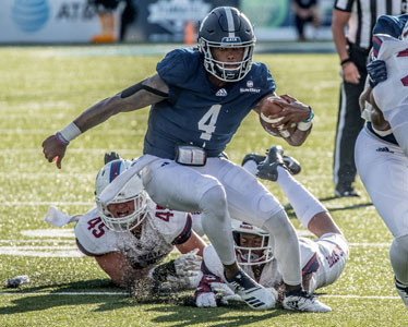 A football player in a navy jersey with the number 4 runs with the ball while being tackled by two opposing players in white uniforms during a game on a sunlit field.