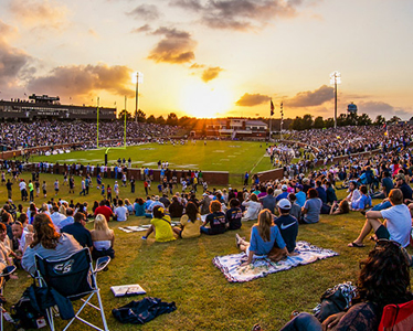Crowds of people sit on a grassy hill and in stadium seats, watching a football game at sunset under partly cloudy skies. Stadium lights are on, and the field is surrounded by fans.