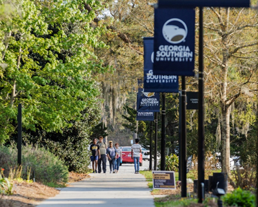 People walk along a tree-lined campus path with several “Georgia Southern University” banners hanging from lampposts. Cars are visible in the background and greenery surrounds the walkway.
