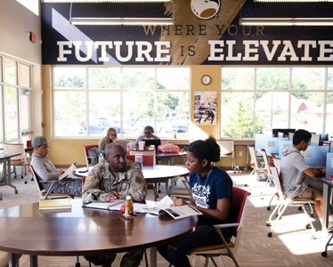 Several people sit studying and talking at tables in a bright, modern room with large windows. A banner on the wall reads, “WHERE YOUR FUTURE IS ELEVATED.” Books and laptops are visible on the tables.