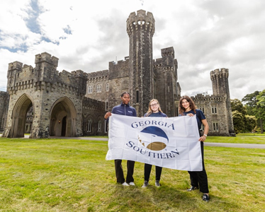 Three people stand on grass in front of a large stone castle, holding a Georgia Southern University flag.
