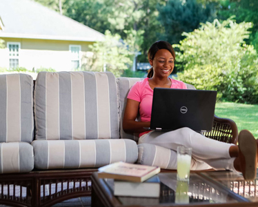 A woman sits on a striped outdoor sofa, smiling while using a laptop. A glass of lemonade and a book rest on a table in front of her. She is on a porch with greenery and a house visible in the background.