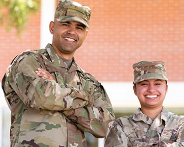 Two people wearing camouflage military uniforms and hats stand side by side outdoors, smiling at the camera, with a brick building and trees in the background.