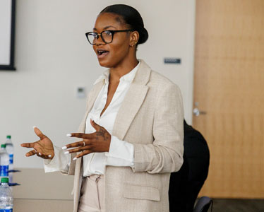 A woman wearing glasses and a beige suit jacket speaks while standing in a meeting room. She gestures with her hands, and a water bottle is visible on the table beside her.