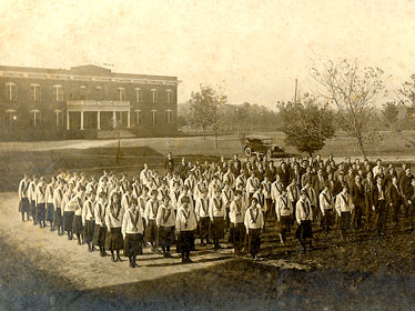 A black-and-white photo shows a large group of students in uniform standing in rows outside a brick school building, with several adults nearby and an early automobile in the background.
