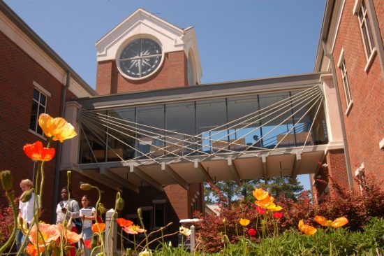 Students walk near the glass bridge and clock tower at the Science Building, with bright flowers in the foreground.