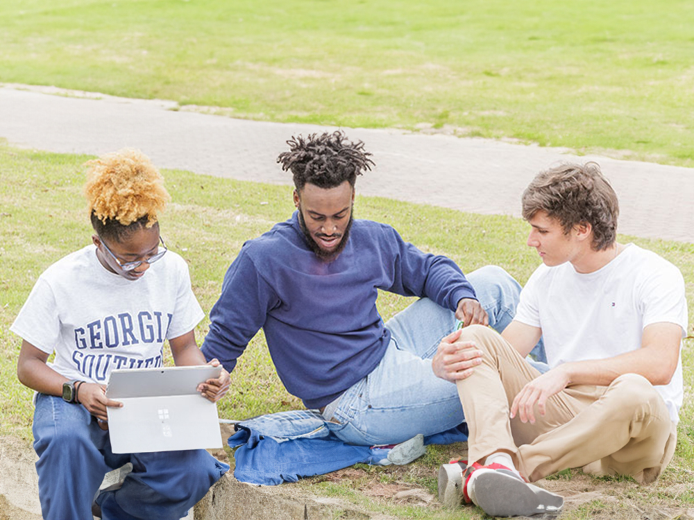 Three college students sitting on grass studying together, one using a tablet and wearing a Georgia Southern t-shirt.