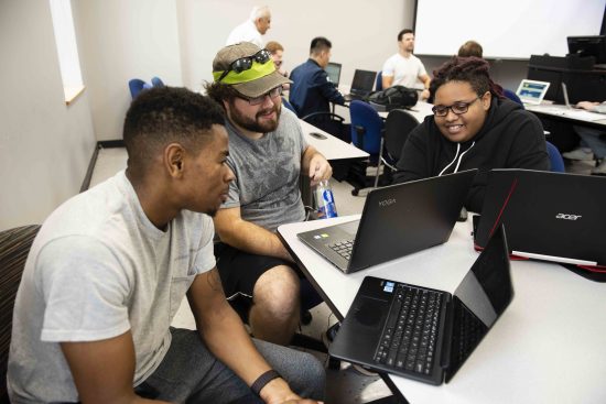 Three students working together on laptops in a classroom during a group project or collaborative learning session.