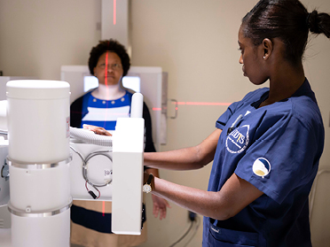 A healthcare worker in blue scrubs operates an X-ray machine while a patient stands against a panel, alignment lasers visible. The healthcare worker adjusts the equipment, focusing on accurate positioning for the scan.