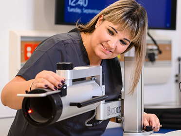 A healthcare professional in scrubs adjusts a Biodex medical imaging device in a clinical setting, with a digital clock and monitor visible in the background.
