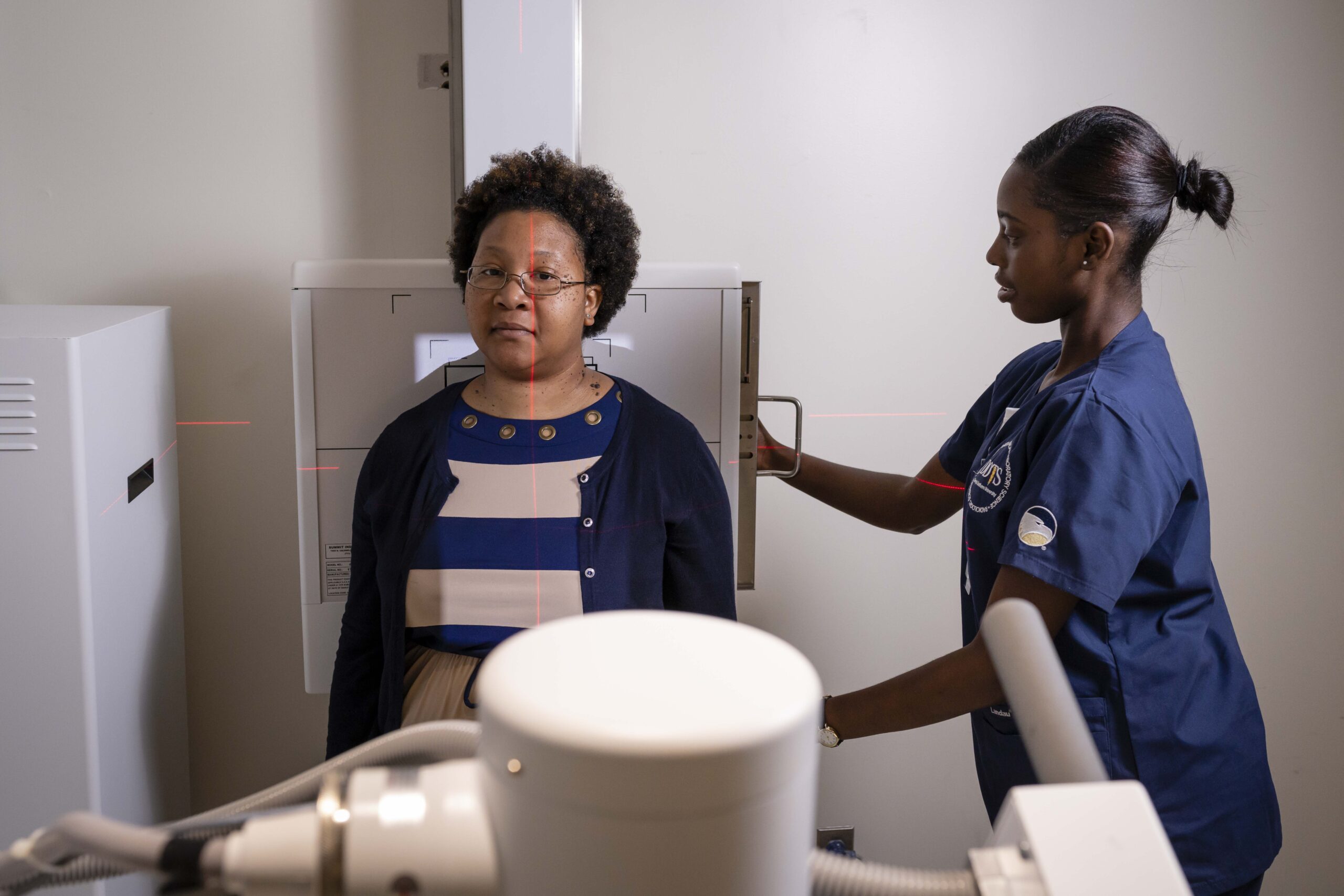 A healthcare worker in blue scrubs prepares a woman for a chest X-ray, positioning her in front of an X-ray machine in a clinical setting. Red laser alignment lines are visible on the wall and the patient.