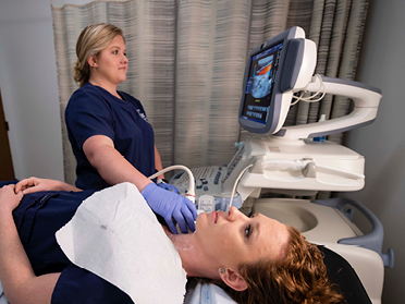 A healthcare professional performs an ultrasound on a patients neck as the patient lies on an exam table, with medical equipment and a monitor nearby.