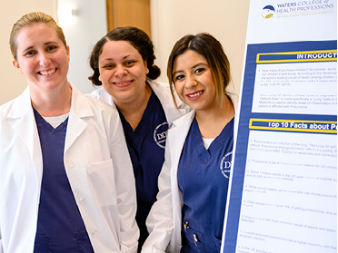 Three women wearing navy scrubs and white lab coats stand smiling next to a presentation board with text. The setting appears to be a professional or academic environment.