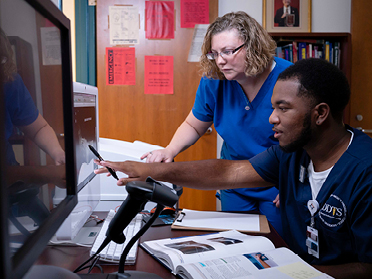 Two healthcare professionals in blue scrubs work together at a desk with a computer, books, and papers; one is pointing at the monitor while the other looks on attentively.