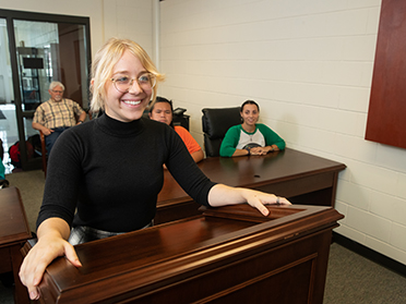 A smiling young woman stands at a wooden podium in a classroom, while three people sit at desks behind her, listening attentively.