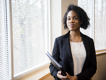 A woman in business attire holds documents and looks thoughtfully out a window with blinds, standing in a bright, modern office space.