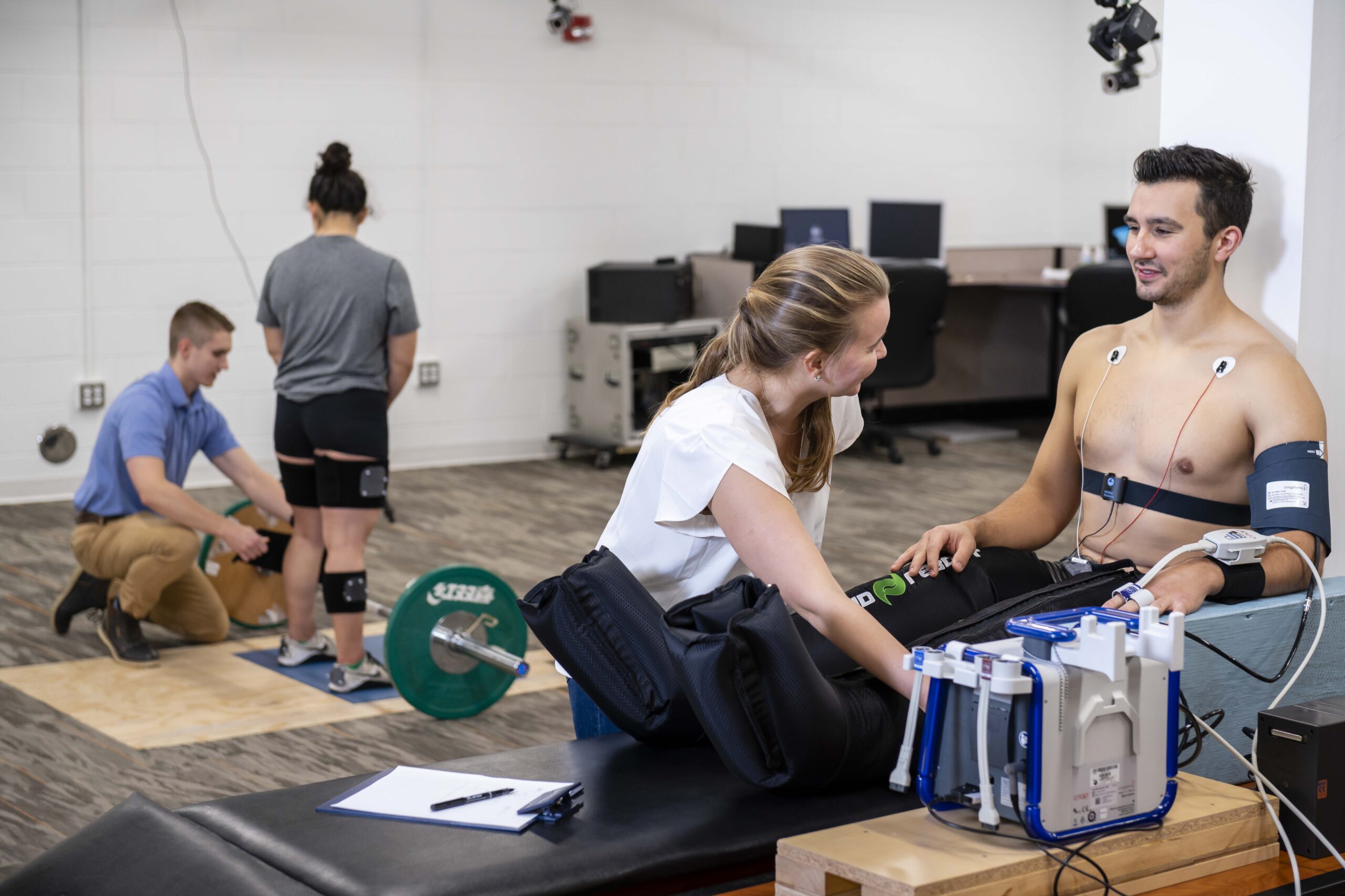 Two people test weightlifting in the background while a woman in the foreground attaches medical sensors to a man seated on a bench, monitoring his physical responses with medical equipment.