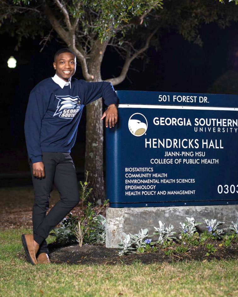 A smiling person in a Georgia Southern University sweatshirt stands next to a blue sign for Hendricks Hall, College of Public Health, at night with trees and grass in the background.