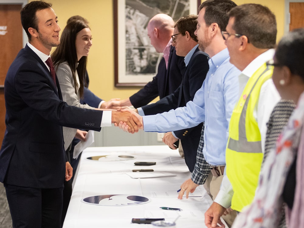 Several people in business attire and one in a safety vest shake hands across a table, appearing to greet or introduce themselves at a professional event or meeting.