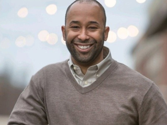 A smiling man with short hair and a beard wears a light-colored collared shirt under a brown sweater. The background is blurry with soft lights and outdoor scenery.